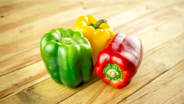 Tres pimientos de colores (rojo, verde, amarillo) sobre una mesa de madera. Los perros pueden comer pimiento.